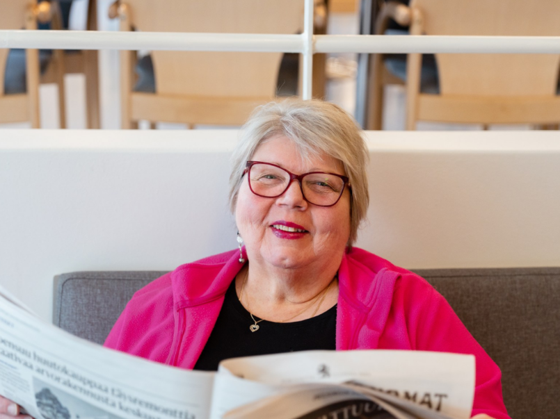 Iäkäs nainen istuu sohvalla ja lukee sanomalehteä. Hän hymyilee ja käyttää punaisia silmälaseja sekä kirkkaan pinkkiä fleecetakkia. Taustalla näkyy vaaleita puutuoleja ja valkoinen kaide.