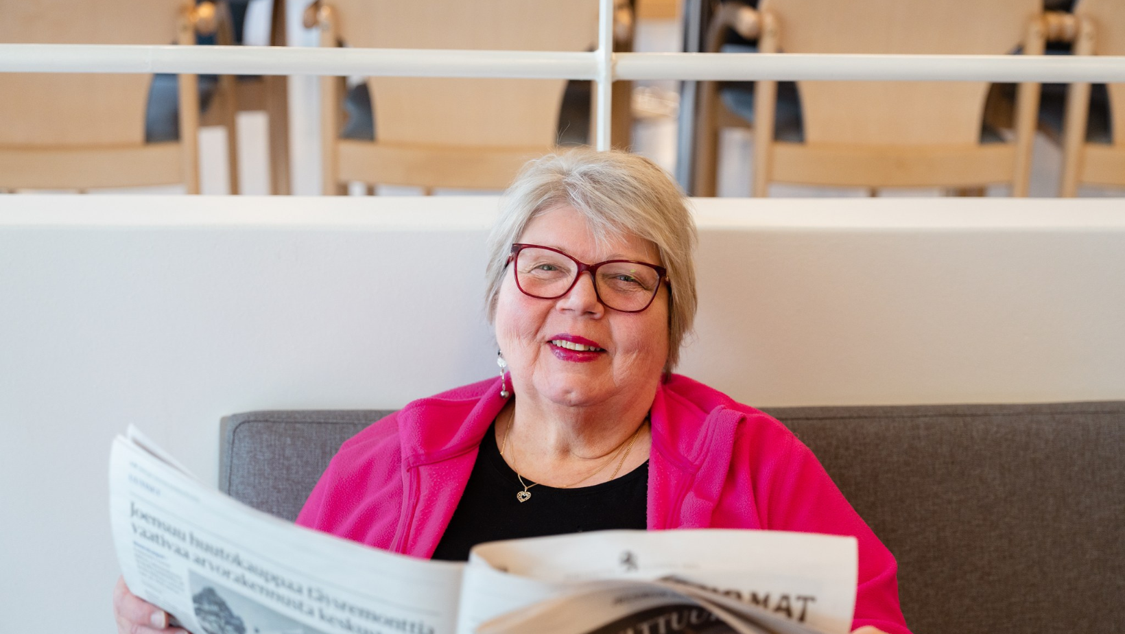 Iäkäs nainen istuu sohvalla ja lukee sanomalehteä. Hän hymyilee ja käyttää punaisia silmälaseja sekä kirkkaan pinkkiä fleecetakkia. Taustalla näkyy vaaleita puutuoleja ja valkoinen kaide.
