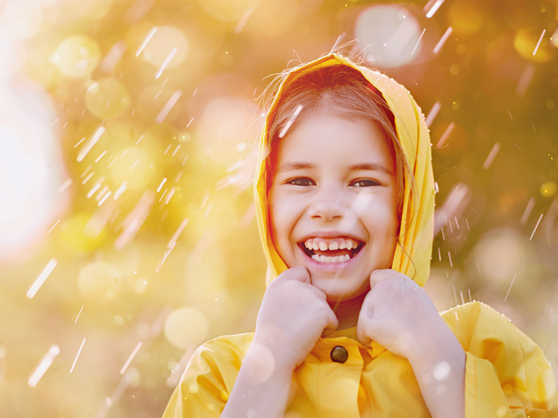 Tyttö seisoo nauraen sateessa keltainen sadetakkipäällään. Taustalla paistaa aurinko.