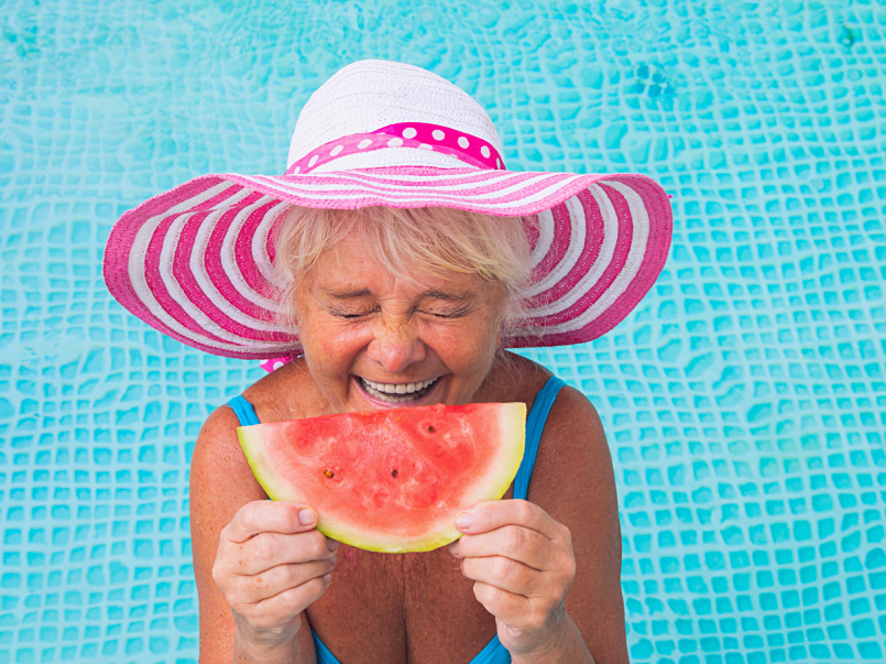 Ikääntynyt nainen seisoo uima-altaassa nauraen ja pitäen kädessään vesimelonin palasta.