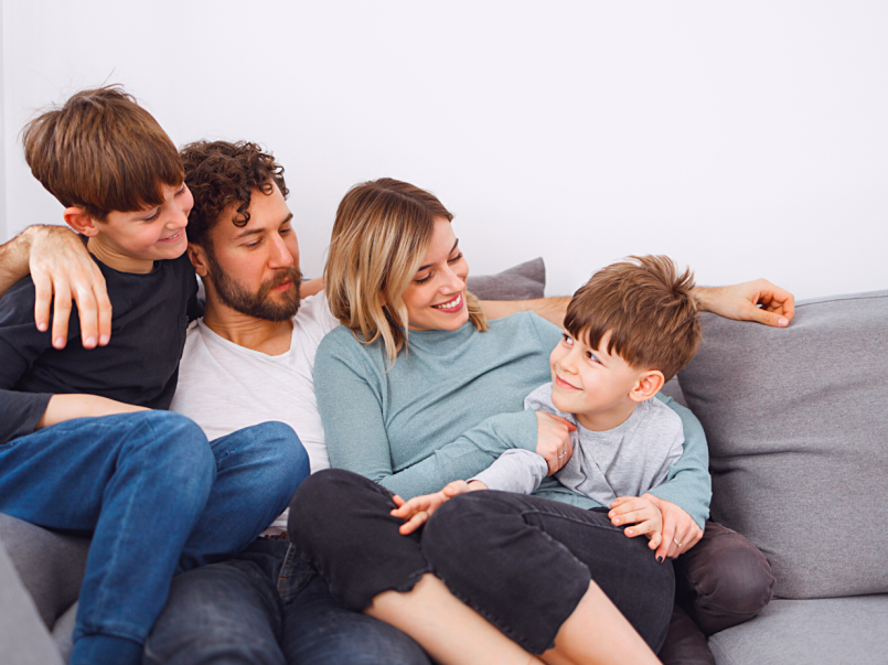 Nelihenkinen perhe istuu hymyillen sohvalla vierekkäin.