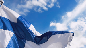 Suomen lippu liehuu tuulessa ja taustalla näkyy sininen taivas ja valkoisia pilviä