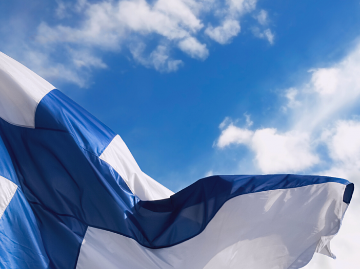 Suomen lippu liehuu tuulessa ja taustalla näkyy sininen taivas ja valkoisia pilviä