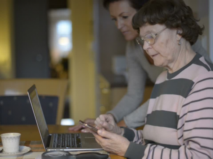Kuvassa ikääntynyt nainen istuu keittiän pyödän äärellä puhelin kädessä ja tietokone edessään, kun taustalla työikäinen nainen auttaa häntä tietokoneen käytössä. Taustalla näkyy sumeana keittiötä ja muuta asuntoa.