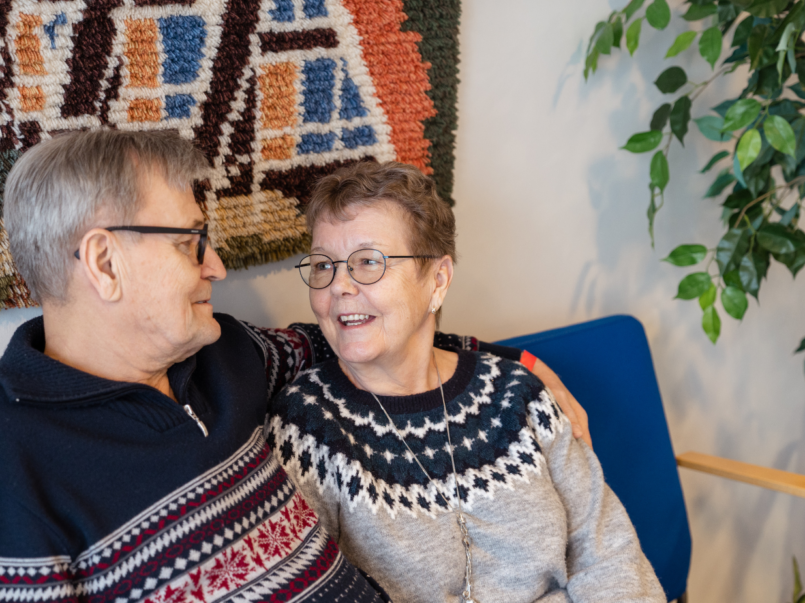 Vanhempi pariskunta istuu sinisellä penkillä hymyillen ja katsoen toisiaan lämpimästi. Molemmilla on yllään kirjoneuleet. Taustalla näkyy värikäs seinävaate ja vihreä huonekasvi.