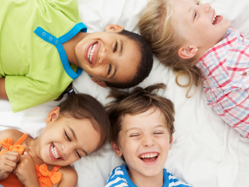 Neljä lasta makaavat sängyssä ja nauravat värikkäissä vaatteissa.