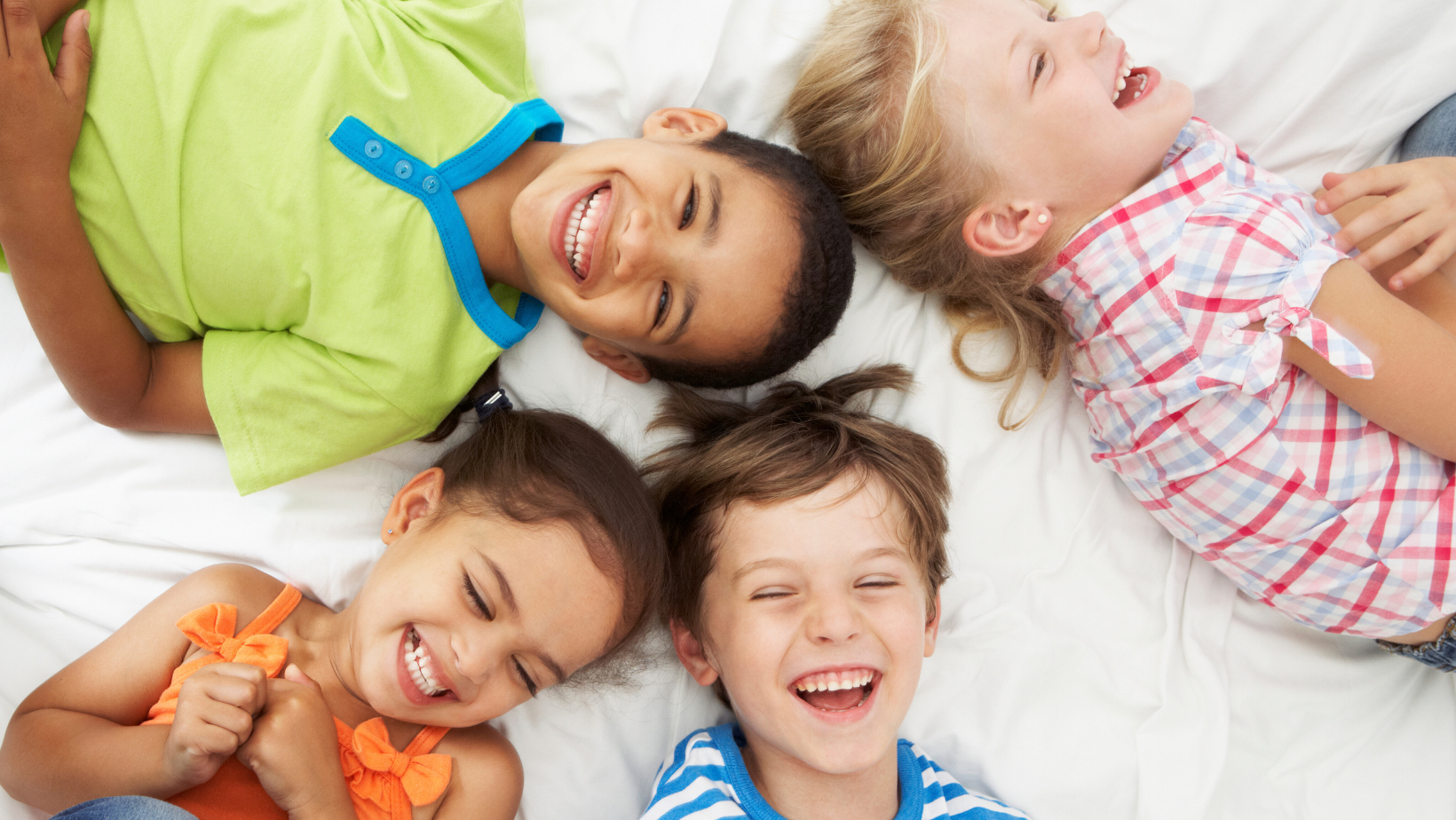Neljä lasta makaavat sängyssä ja nauravat värikkäissä vaatteissa.