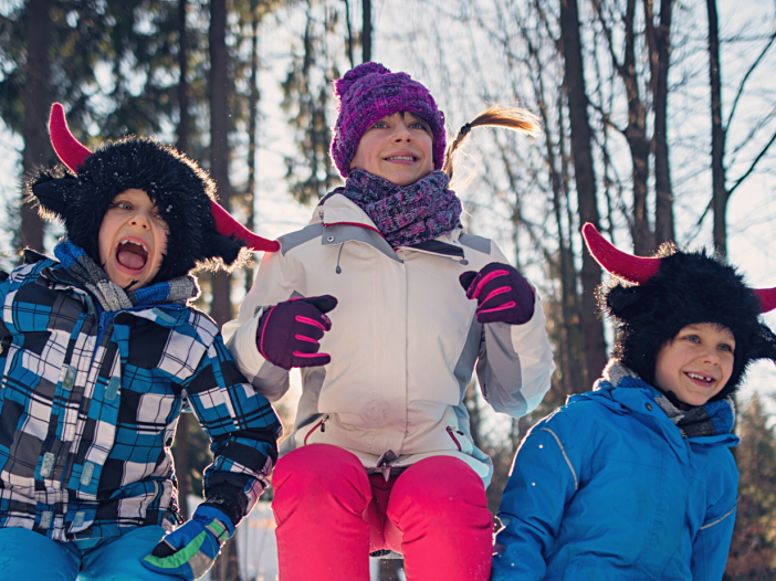 Äiti ja kaksi kouluikäistä lasta hyppää ilmaan iloiset ilmeet kasvoillaan ja värikkäät talvivaatteet päällään. Takana näkyy talvinen metsä.