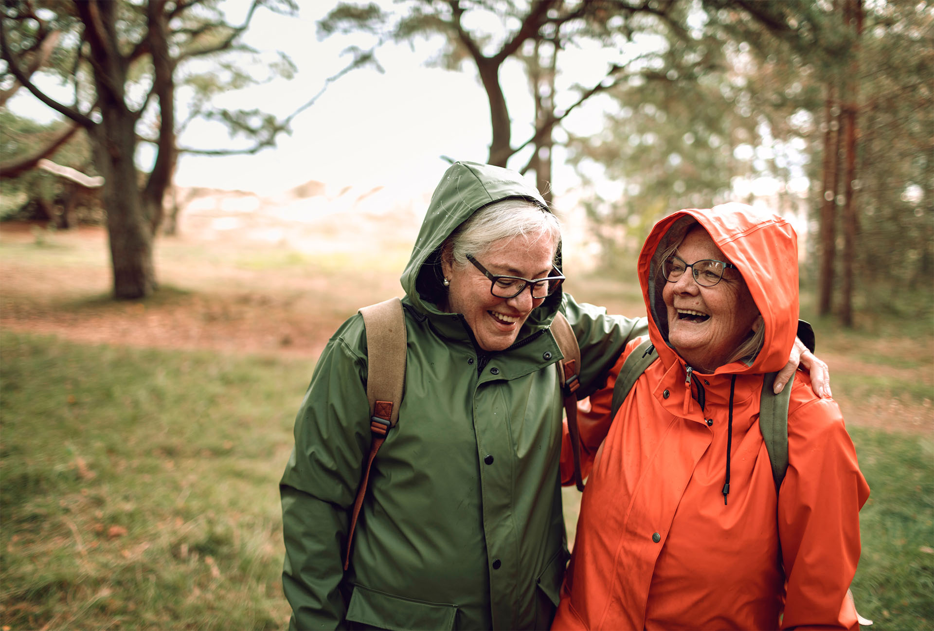 Kaksi ikääntynyttä naista pitää toisiaan olkapäistä kiinni nauraen. Heillä on sadetakit päällä syksyisessä metsämaisemassa.