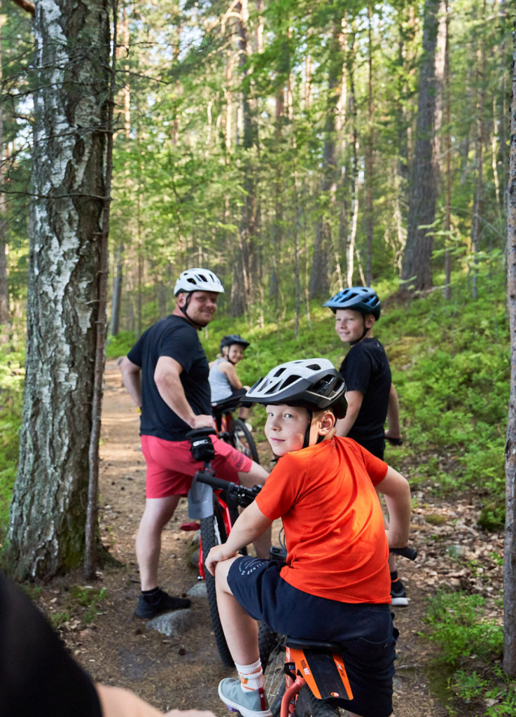 Isä ja kolme lasta ovat kääntyneet katsomaan kohti kameraa pyörälenkillä metsässä.