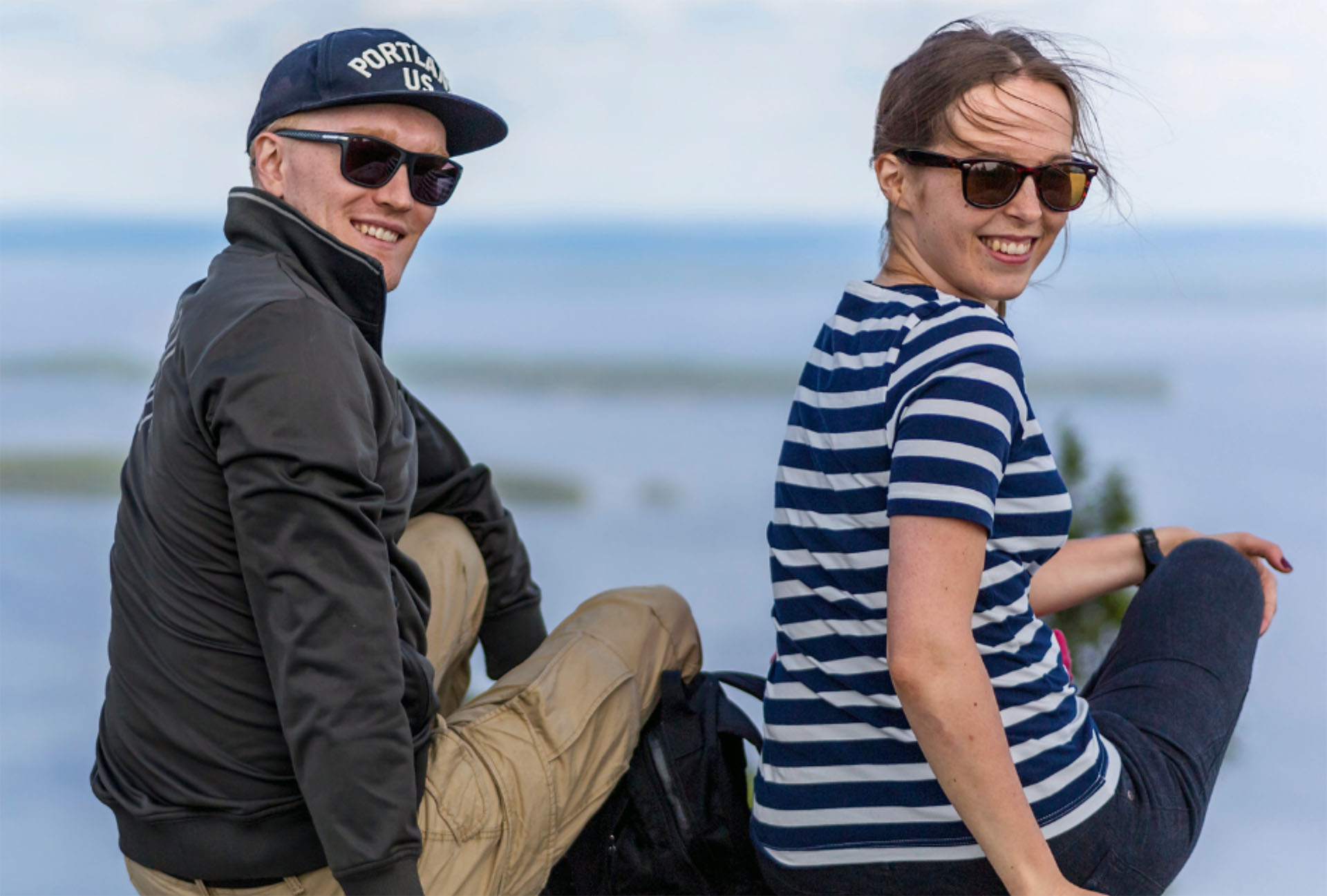 Mies ja nainen istuvat kalliolla saaristossa. He ovat kääntyneet kohti kameraa ja hymyilevät aurinkolasit päässään.